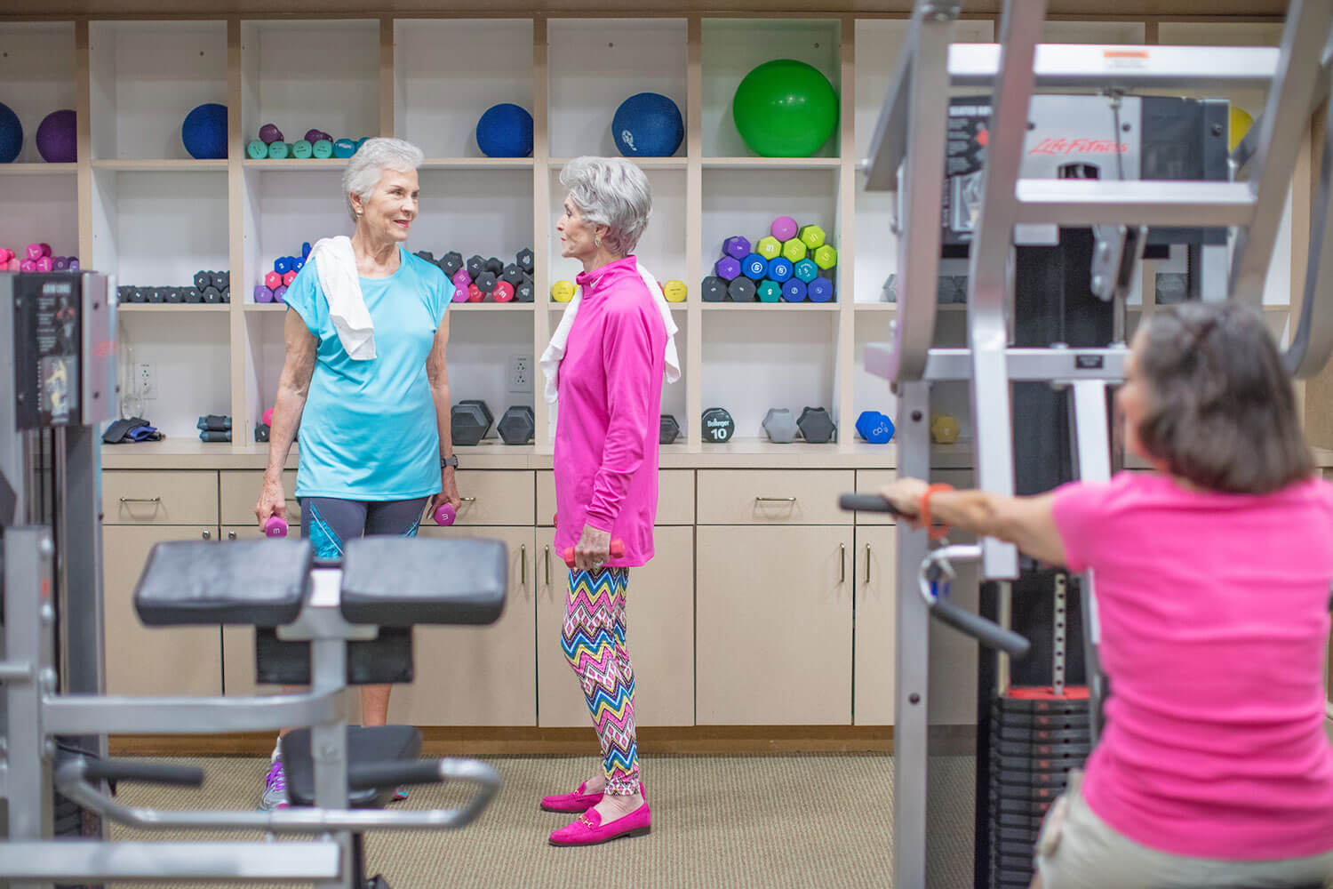 Senior women standing together in a fitness center dressed in workout clothing & holding small weights