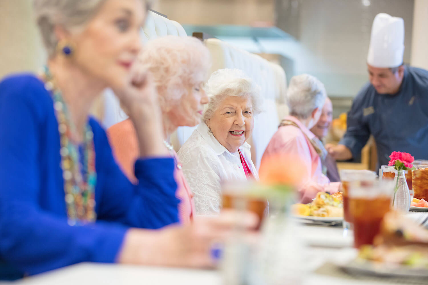 Senior woman seated at a long dining table conversing as they wait for their food