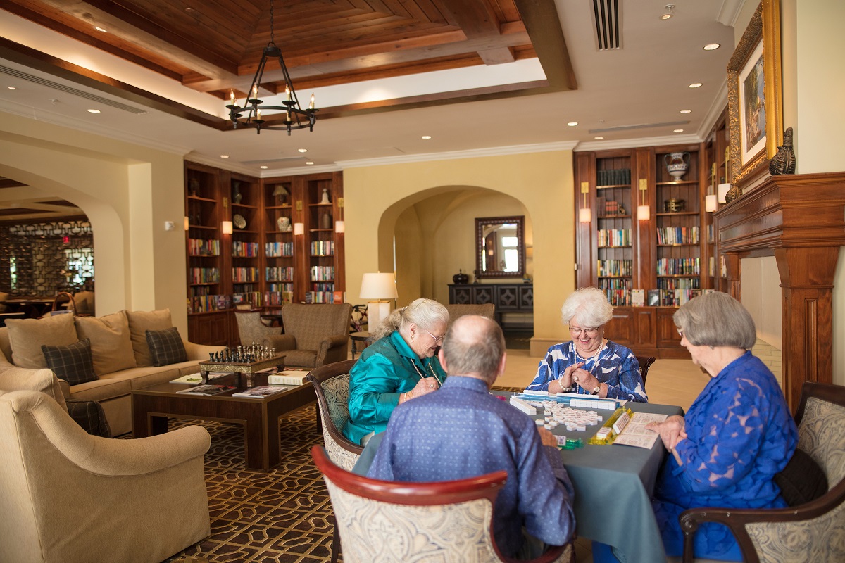 Seniors seated in an elegant game room playing dominos together surrounded by tall bookshelves filled with books