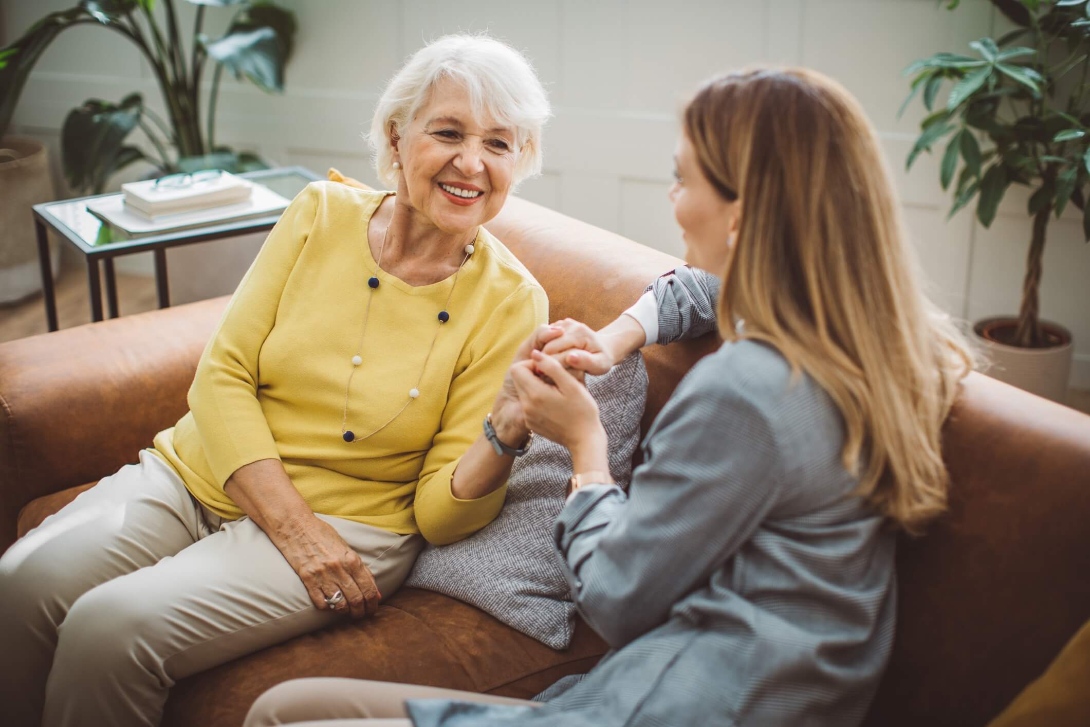 Senior woman sitting on couch with adult daughter holding hands, smiling, & talking