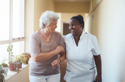 Older woman with young nurse woman holding hands & laughing as they walk down a brightly lit corridor