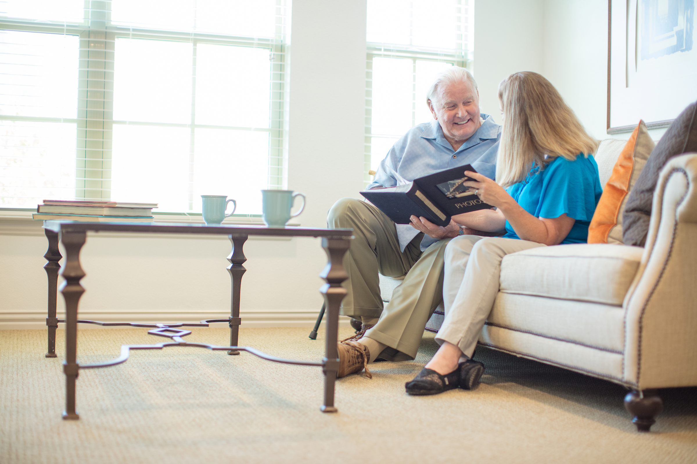 Older adult gentleman sitting on a couch smiling while looking at a photo album with his adult daughter