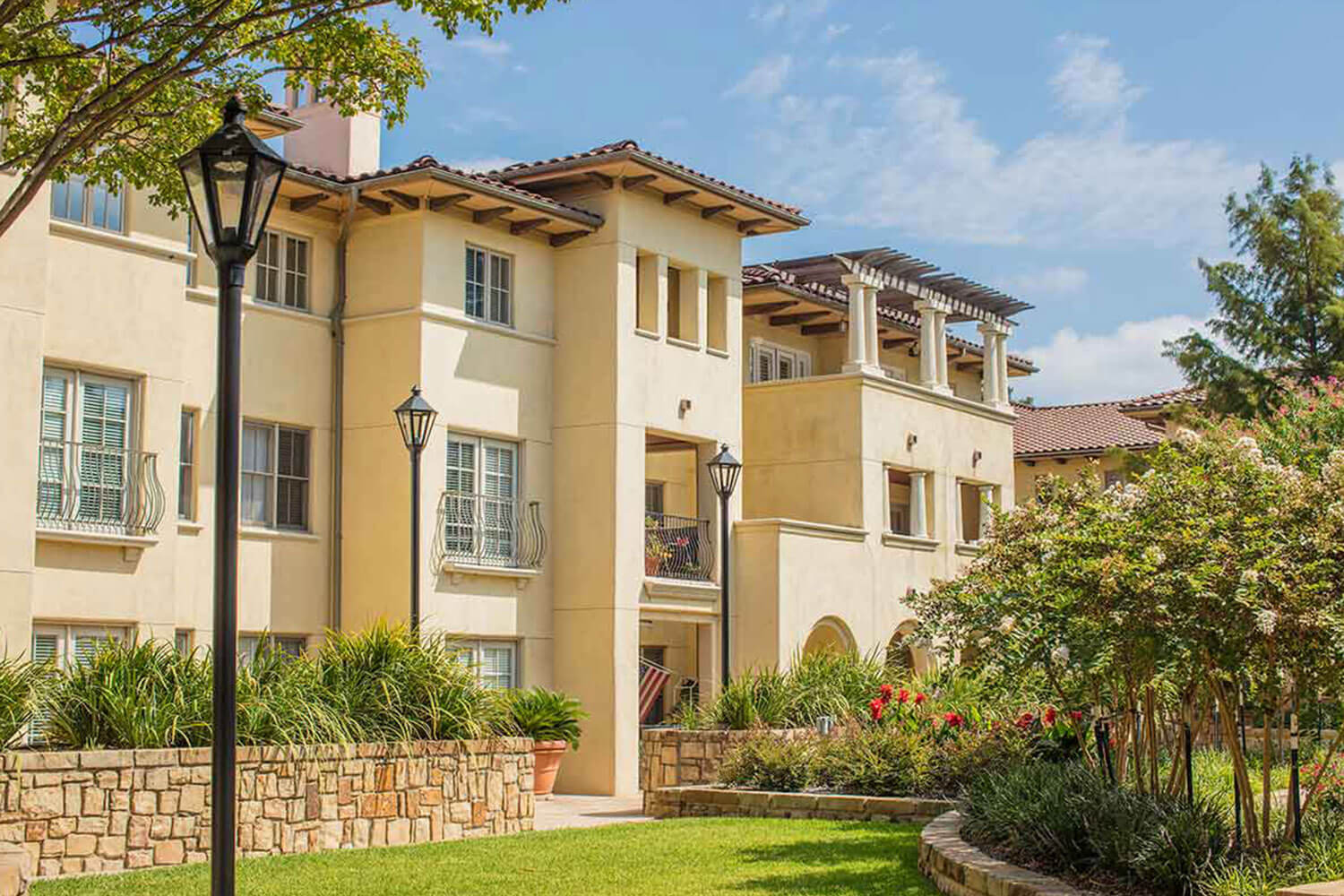 Large 3 story off white stucco senior living apartment building with beautifully landscaped courtyard with raised flowerbeds full of flora