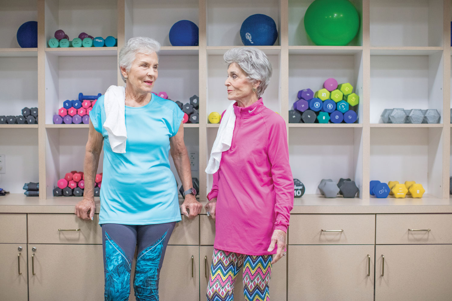 Two mature adult women standing & chatting together in front of shelving with workout equipment & barbells