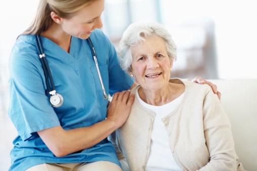 Close up of nurse in blue shirt with her hands on a smiling & seated senior woman with white hair